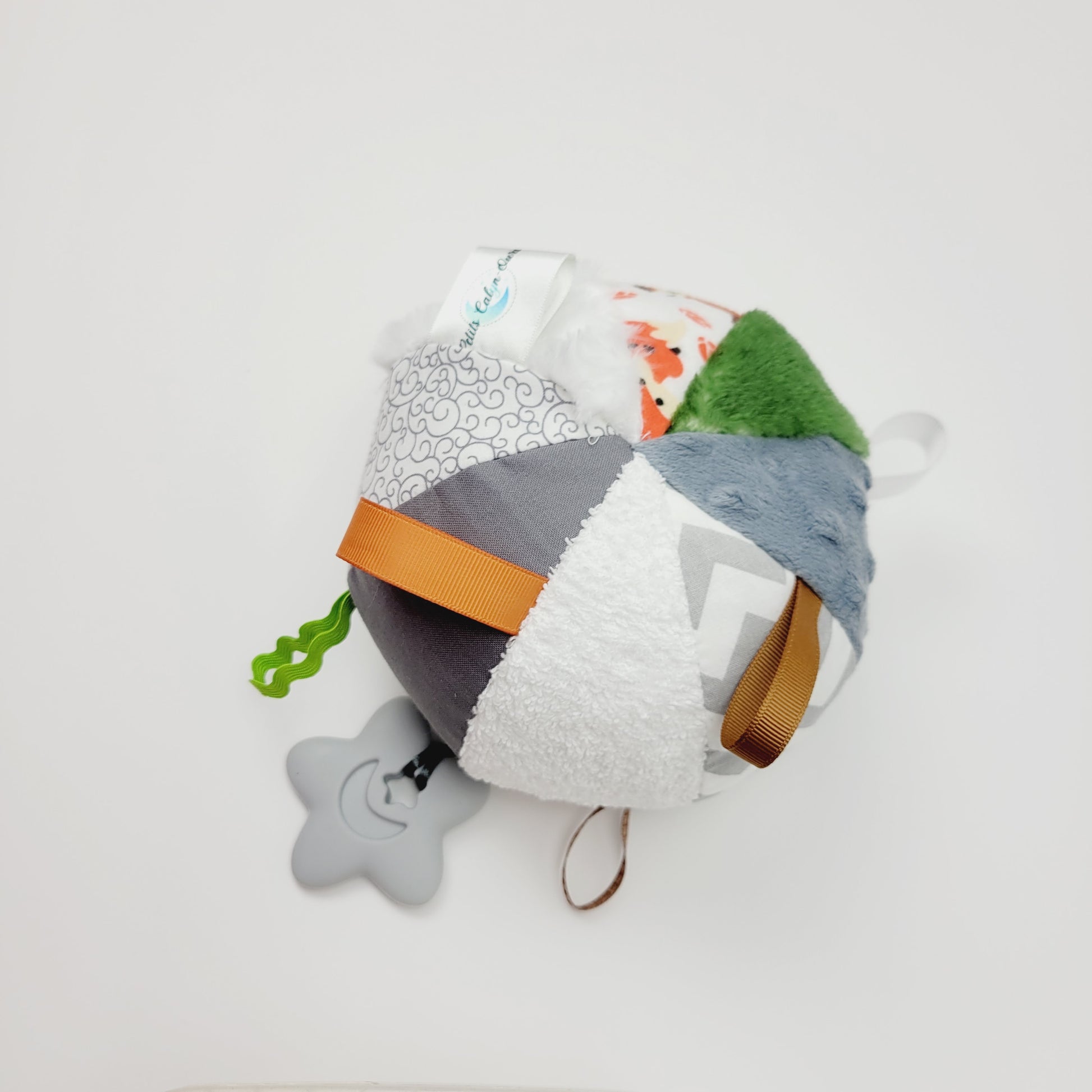 Ballon d'éveil pour bébé avec 8 tissus différents pour créer de la texture. On y retrouve un polar vert, du polar à pois gris, de la flanelle à chevron blanc et gris, de la ratine blanche, du coton blanc et gris, de la rosette blanche, de la flanelle imprimé avec des renards et des hiboux. Entre les différent tissu, il y a des petits rubans vert, orange, noir, blanc, brun et au bout d'un de ces rubans y est attaché un jouet de dentition en silicone gris en forme d'étoile avec une Lune à l'intérieur