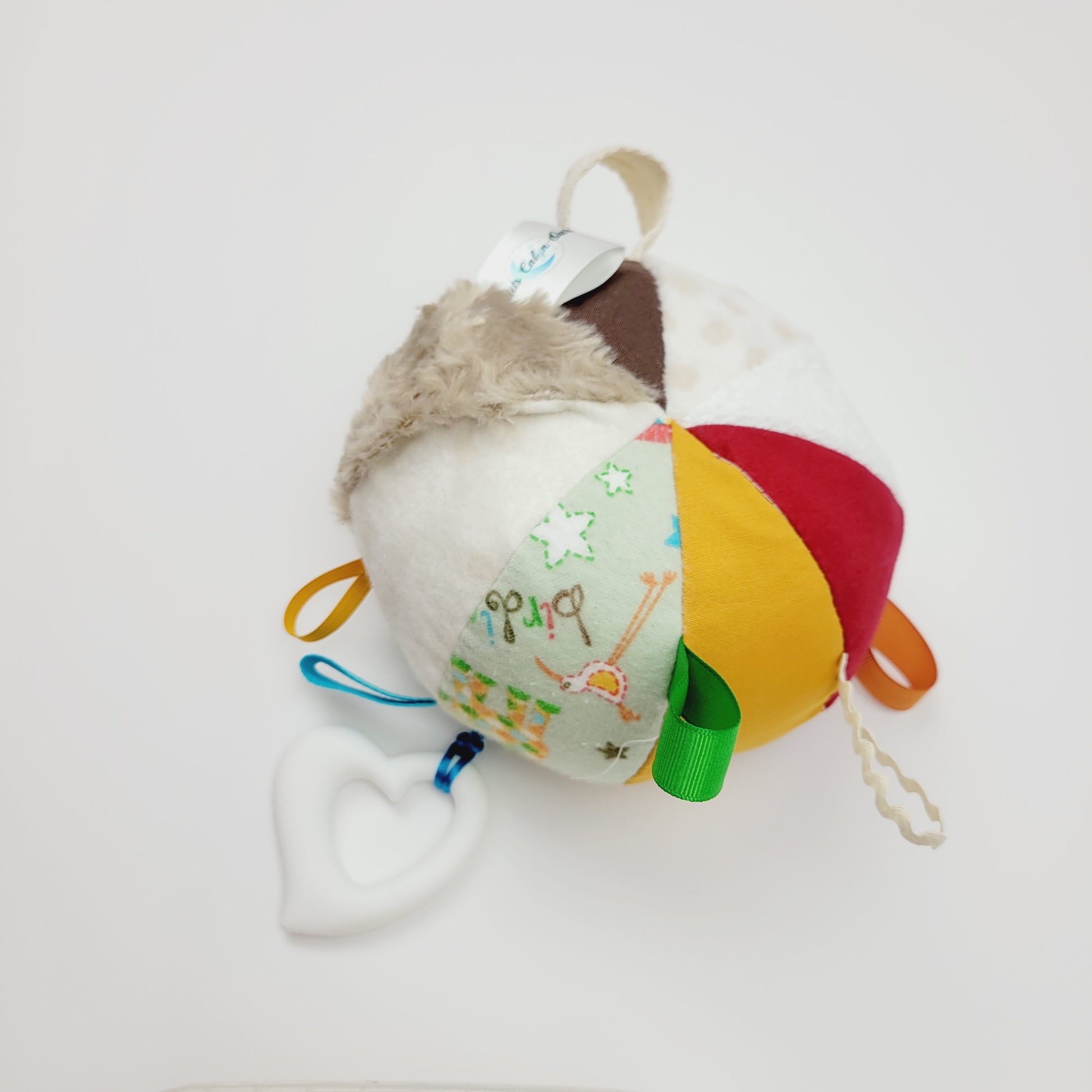 Ballon d'éveil pour bébé de 3-9 mois. Le ballon est composé de 8 différents tissus pour les différentes textures. Sur ce ballon nous retrouvons une ratine blanche, du corde-du-roi rouge et jaune et brun, une flanelle imprimé avec des girafes, un polar blanc et un autre beige, une rosette beige. Entre les tissus ont retrouve des petits rubans de couleur brun, jaune, vert, bleu et beige. Au bout de l'un de ses rubans y est attaché un jouet de dentition en silicone en forme de coeur de couleur blanc.