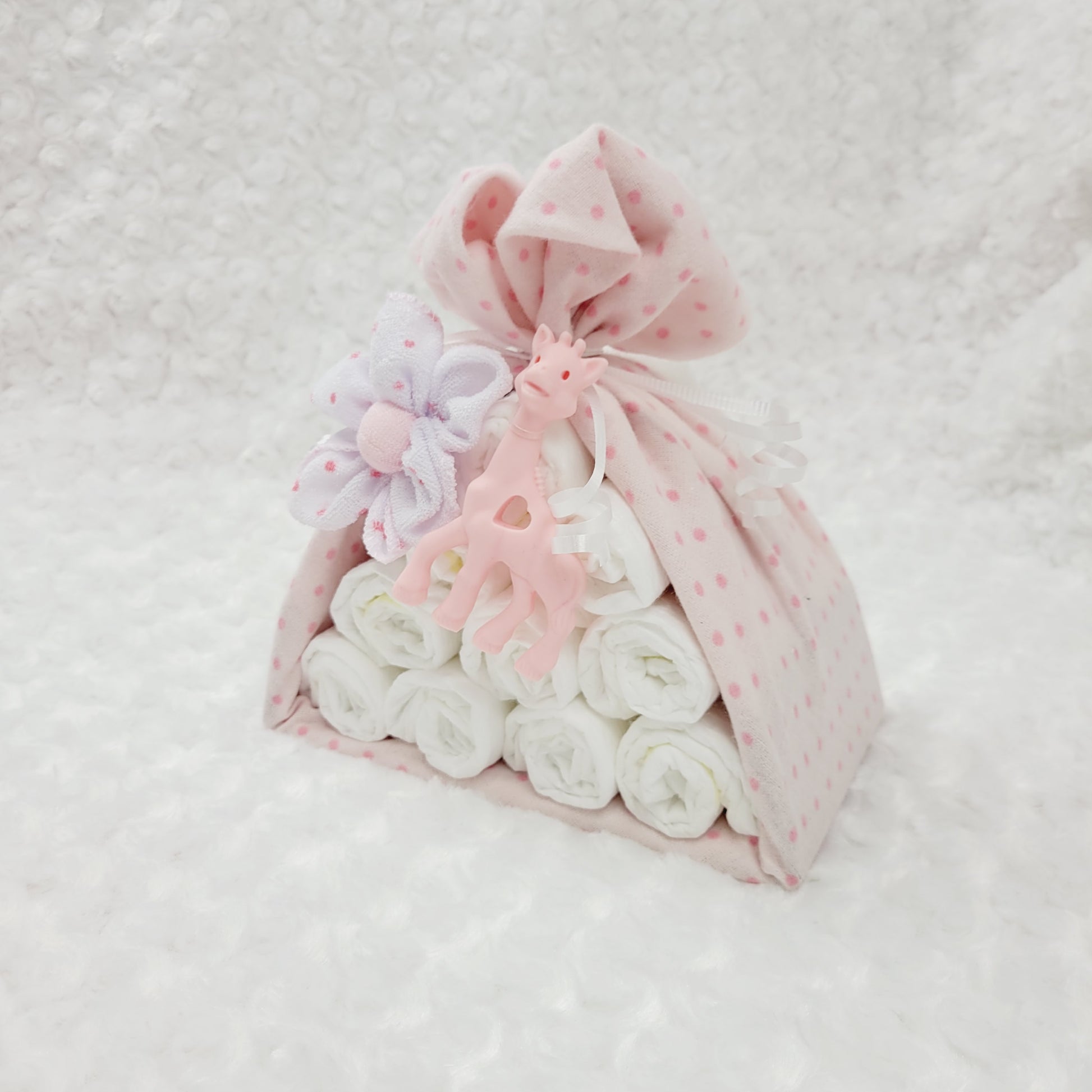 Ensemble cadeau baluchon avec une couverture en flanelle rose à pois. Contient 10 couches pour bébé de taille #2 ainsi que 2 débarbouillettes en disposées en fleur. Complété d'un jouet de dentition en silicone en forme de girafe rose.