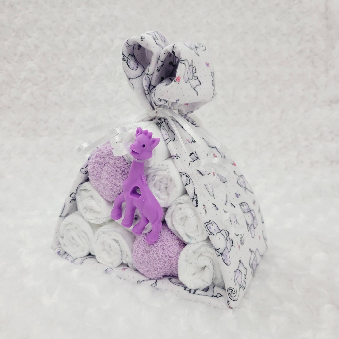Ensemble cadeau baluchon avec une couverture de flanelle blanche avec des petits éléphant mauves. Contient 10 couche pour bébé de taille #2 une paire de bas chaud de couleur mauve. Complété d'un jouet de dentition en silicone en forme de girafe mauve.