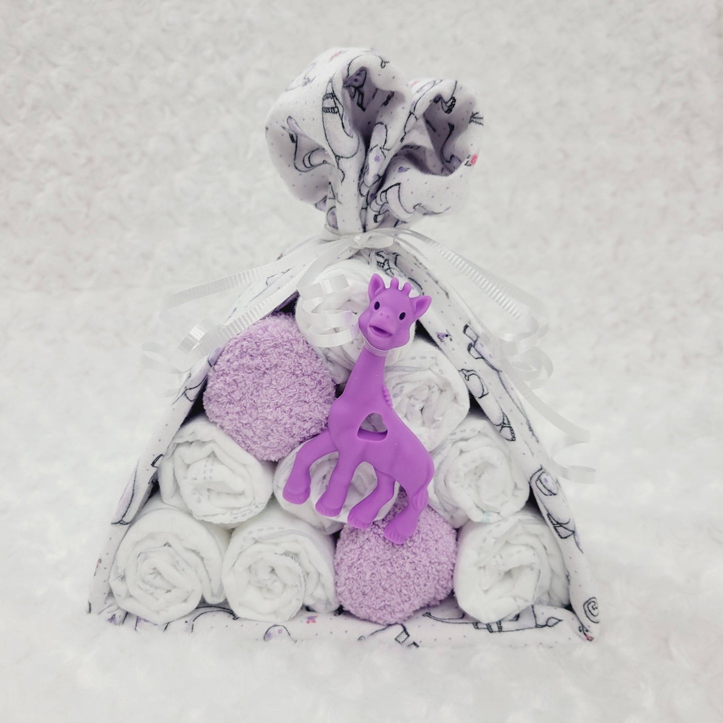 Ensemble cadeau en forme de baluchon avec une couverture blanche avec des petits éléphant mauve. Contient 10 couche pour bébé de taille #2 avec une paire de bas chaud de couleur mauve. Complété d'un jouet de dentition en silicone en forme de girafe mauve.