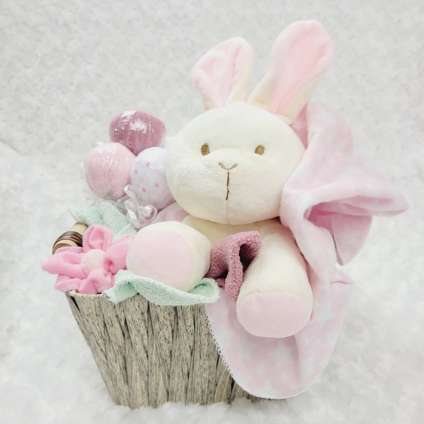 De face, Ensemble cadeau pour babyshower dans un panier brun contenant des couches pour bébé de taille #2 des débarbouillettes disposé en suçon, un toutou lapin beige, une couverture en flanelle rose et des bas chaud.