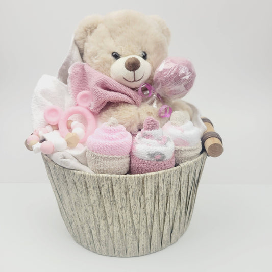 Ensemble cadeau pour babyshower de bébé fille. Un panier contenant un toutou ourson beige, une couverture en flanelle des débarbouillettes disposées en cupecake et des bas en forme de suçon. Complété d'un jouet de dentition en silicone.