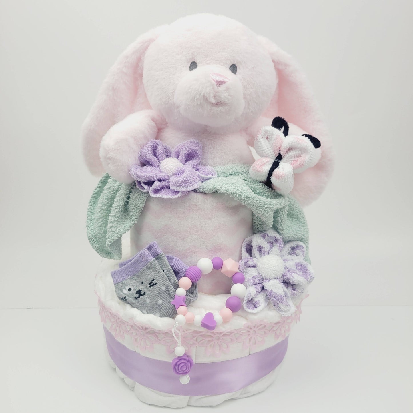 Ensemble cadeau pour babyshower de bébé fille. Gâteau 2 étages avec un toutou lapin inséré dans l'étage du dessus. On retrouve des bas sur l'étage du bas ainsi qu'un jouet de dentition en silicone. Trouvez aussi les fleur fait en débarbouillettes et même le papillon.