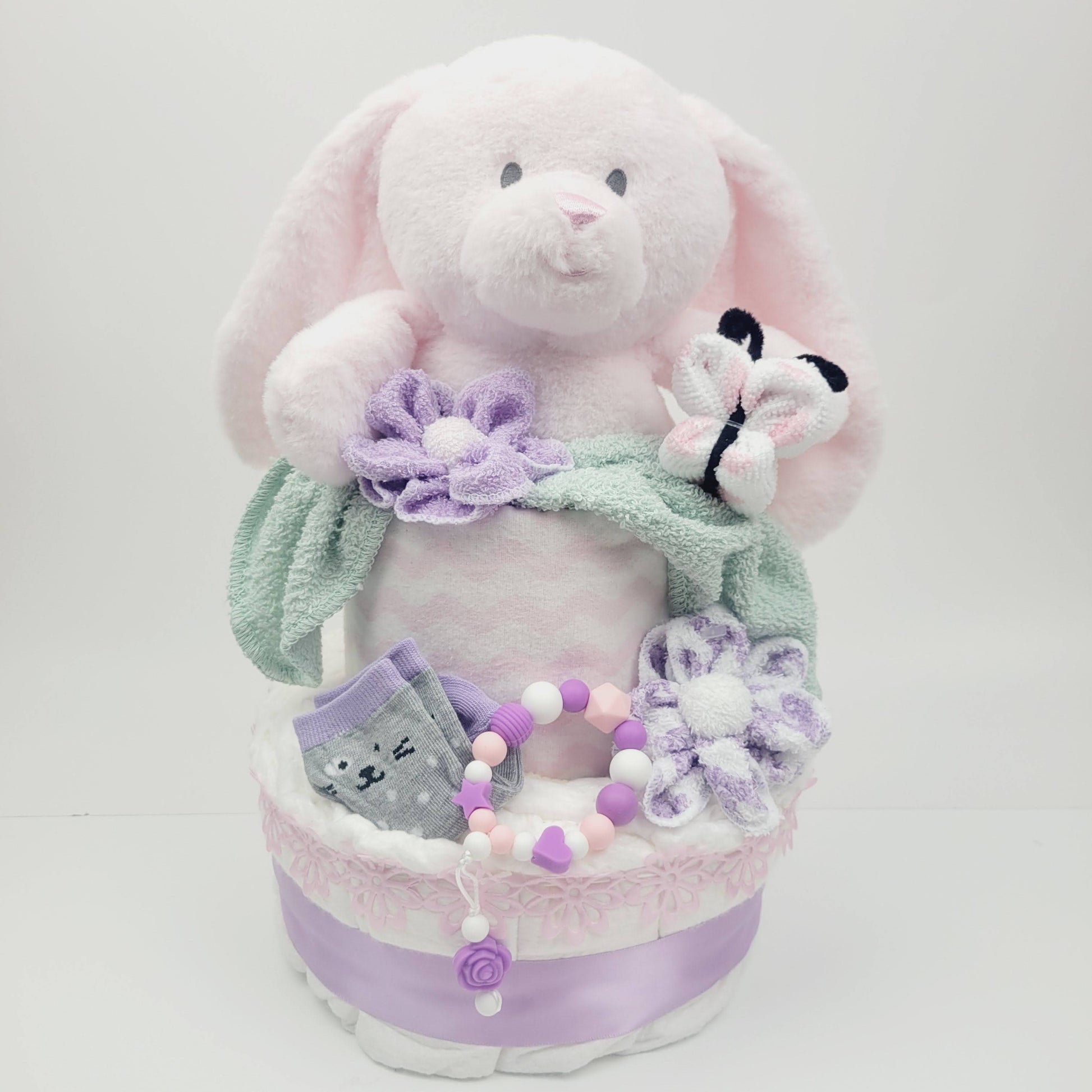 Ensemble cadeau pour babyshower de bébé fille. Gâteau 2 étages avec un toutou lapin inséré dans l'étage du dessus. On retrouve des bas sur l'étage du bas ainsi qu'un jouet de dentition en silicone. Trouvez aussi les fleur fait en débarbouillettes et même le papillon.