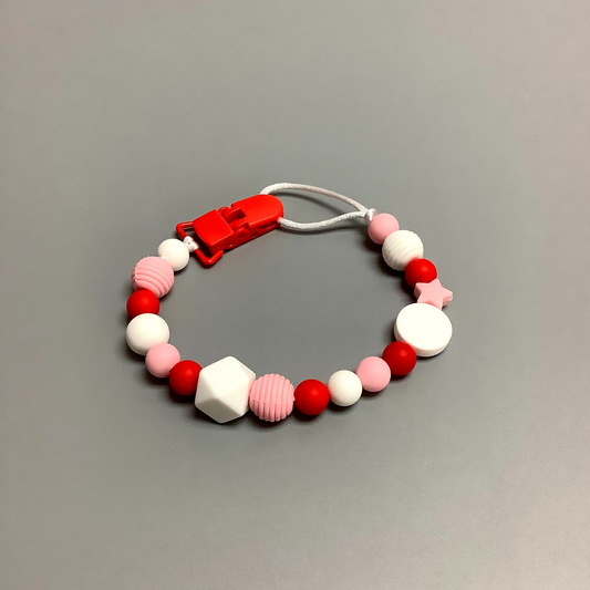 Attache-suce en silicone fait avec plusieurs billes en silicones de différentes couleurs, tailles et formes, et il est complété par une attache en plastique.