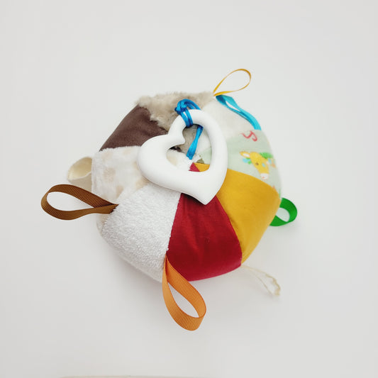 Ballon d'éveil pour bébé de 3-9 mois. Le ballon est composé de 8 différents tissus pour les différentes textures. Sur ce ballon nous retrouvons une ratine blanche, du corde-du-roi rouge et jaune et brun, une flanelle imprimé avec des girafes, un polar blanc et un autre beige, une rosette beige. Entre les tissus ont retrouve des petits rubans de couleur brun, jaune, vert, bleu et beige. Au bout de l'un de ses rubans y est attaché un jouet de dentition en silicone en forme de coeur de couleur blanc.