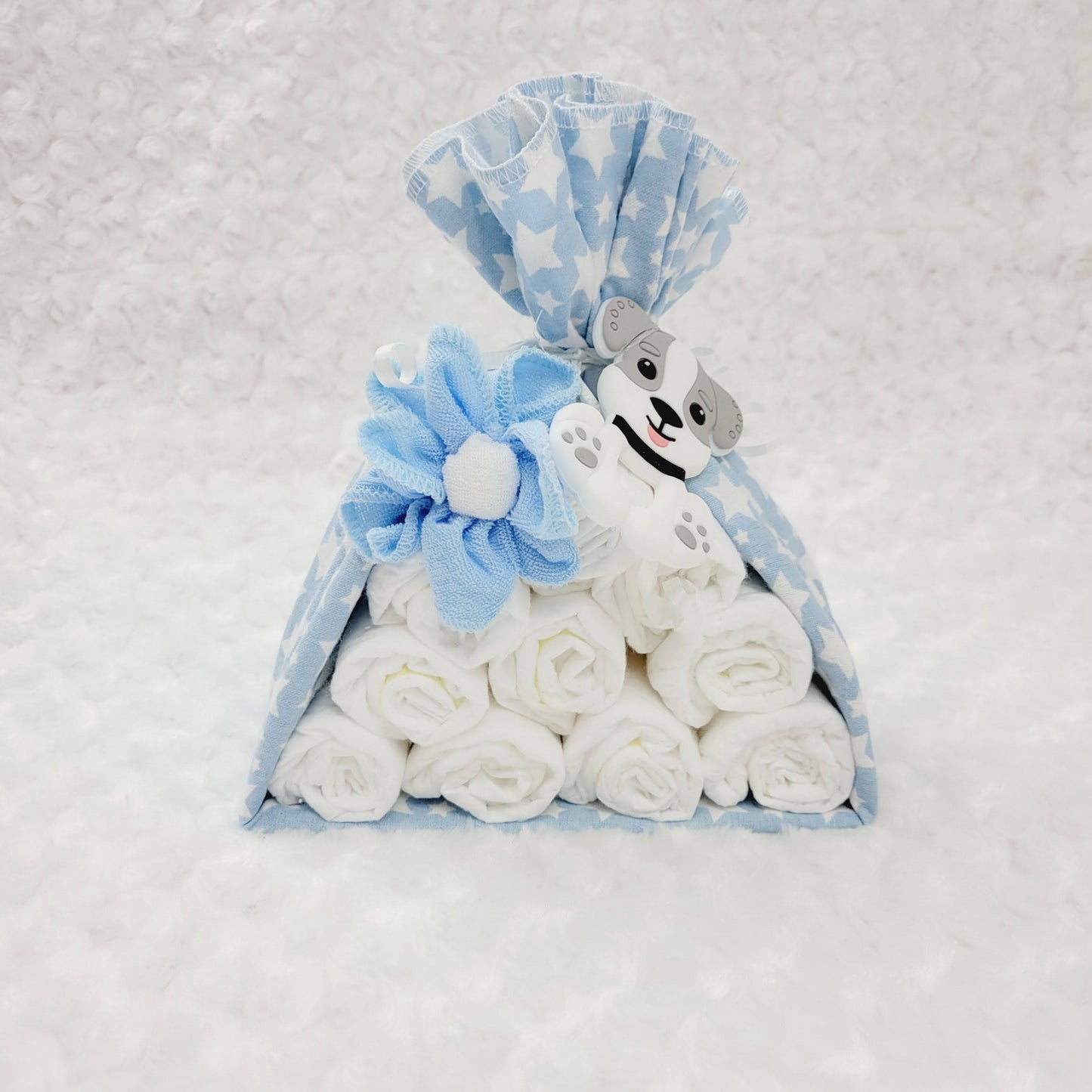 Baluchon avec une couverture bleu étoilé blanc avec 10 couches de taille #2 avec une fleur fait de débarbouillettes bleu et blanche. Complété avec un jouet de dentition en silicone en forme de chien de couleur blanc et gris.