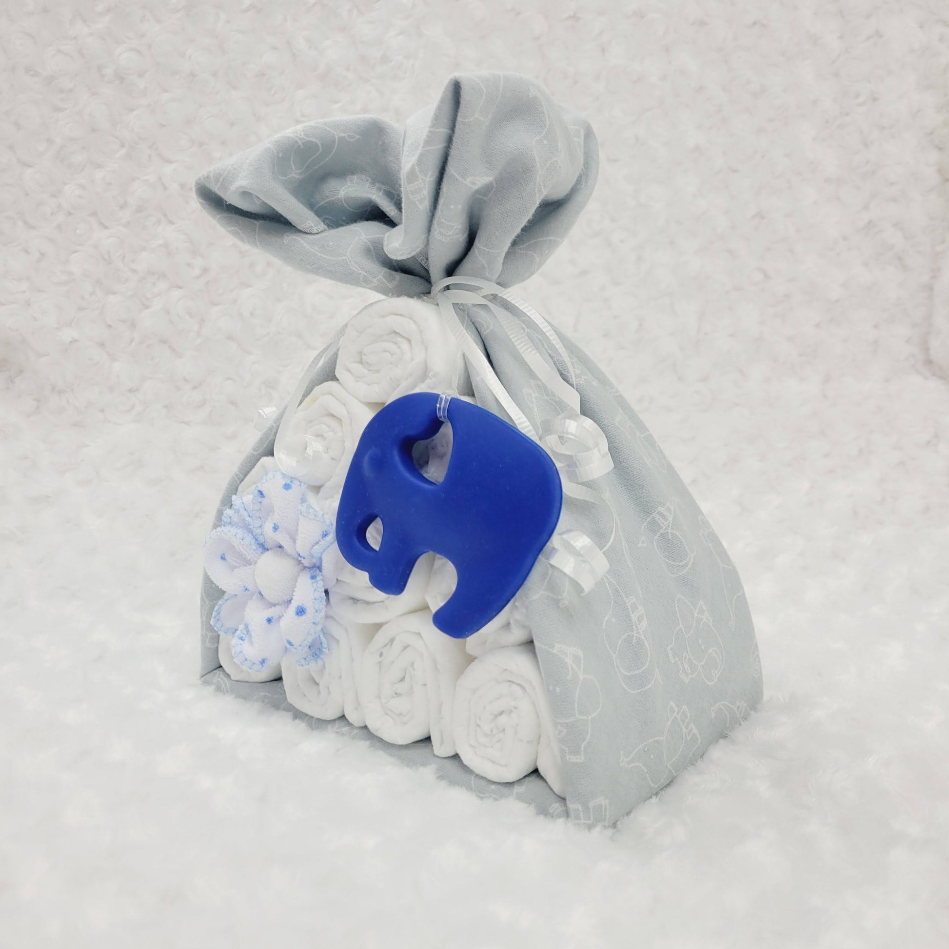 Baluchon avec une couverture gris en flanelle avec un imprimé de petits éléphants blanc. Le baluchon contient 10 couches pour bébé de taille #2 avec 2 débarbouillettes disposé en forme de fleur. Complété d'un jouet de dentition en silicone en forme d'éléphant de couleur bleu royal.
