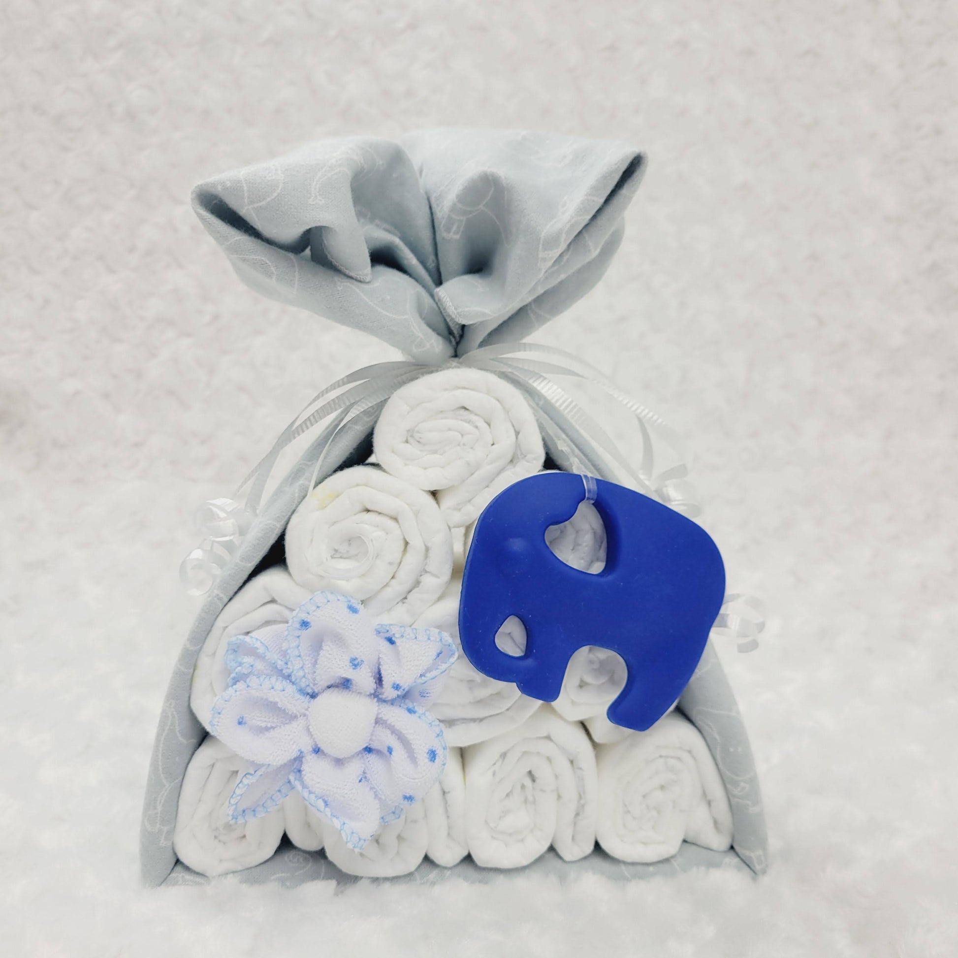 Baluchon vu de face avec un couverture grise imprimé d'éléphant blanc. L'ensemble cadeau contient 10 couches pour bébé de taille #2 et 2 débarbouillettes en forme de fleur. Complété d'un jouet de dentition en silicone en forme d'éléphant bleu royal.