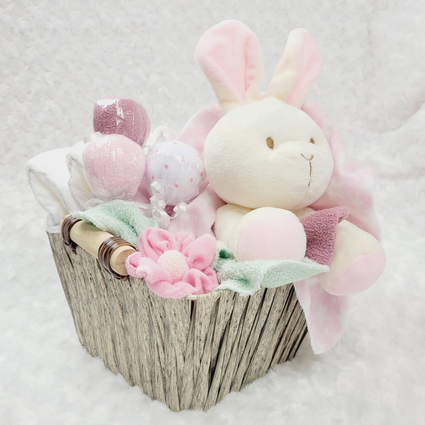 Ensemble cadeau pour babyshower dans un panier brun contenant des couches pour bébé de taille #2 des débarbouillettes disposé en suçon, un toutou lapin beige, une couverture en flanelle rose et des bas chaud. 