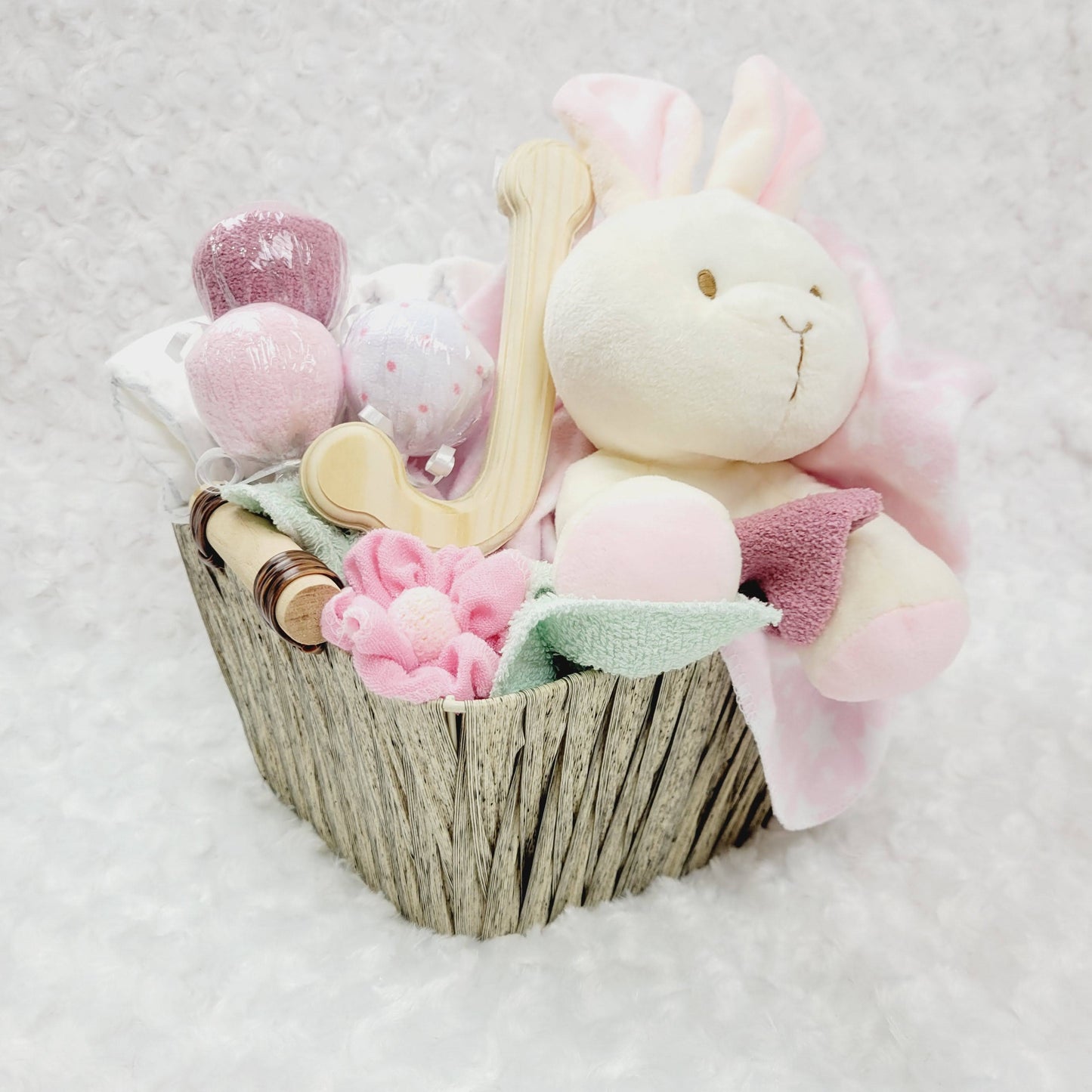 Ensemble cadeau pour babyshower dans un panier brun contenant des couches pour bébé de taille #2 des débarbouillettes disposé en suçon, un toutou lapin beige, une couverture en flanelle rose et des bas chaud. Personnalisé avec la lettre J en bois naturel.