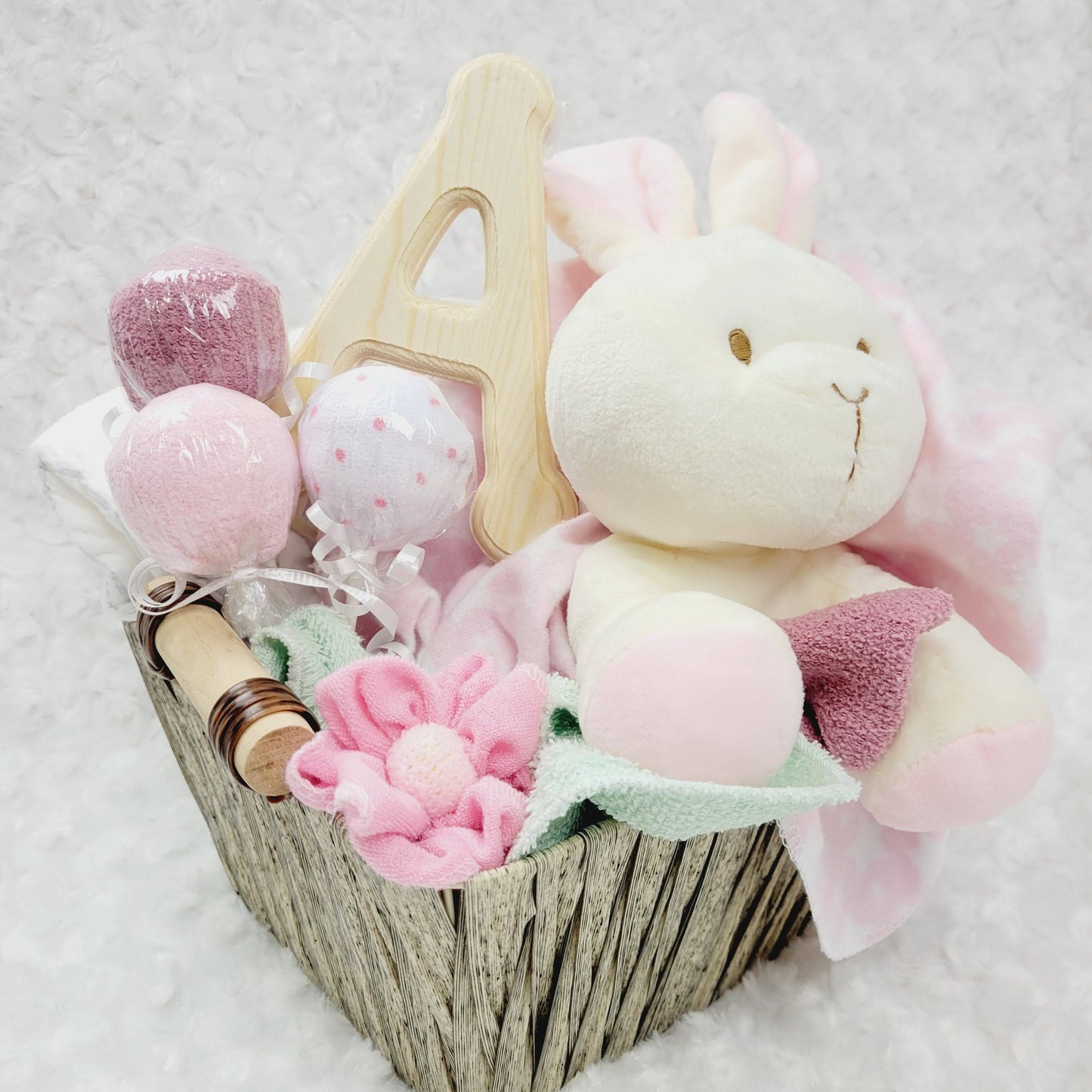 Ensemble cadeau pour babyshower dans un panier brun contenant des couches pour bébé de taille #2 des débarbouillettes disposé en suçon, un toutou lapin beige, une couverture en flanelle rose et des bas chaud. Personnalisé avec la lettre A en bois naturel.