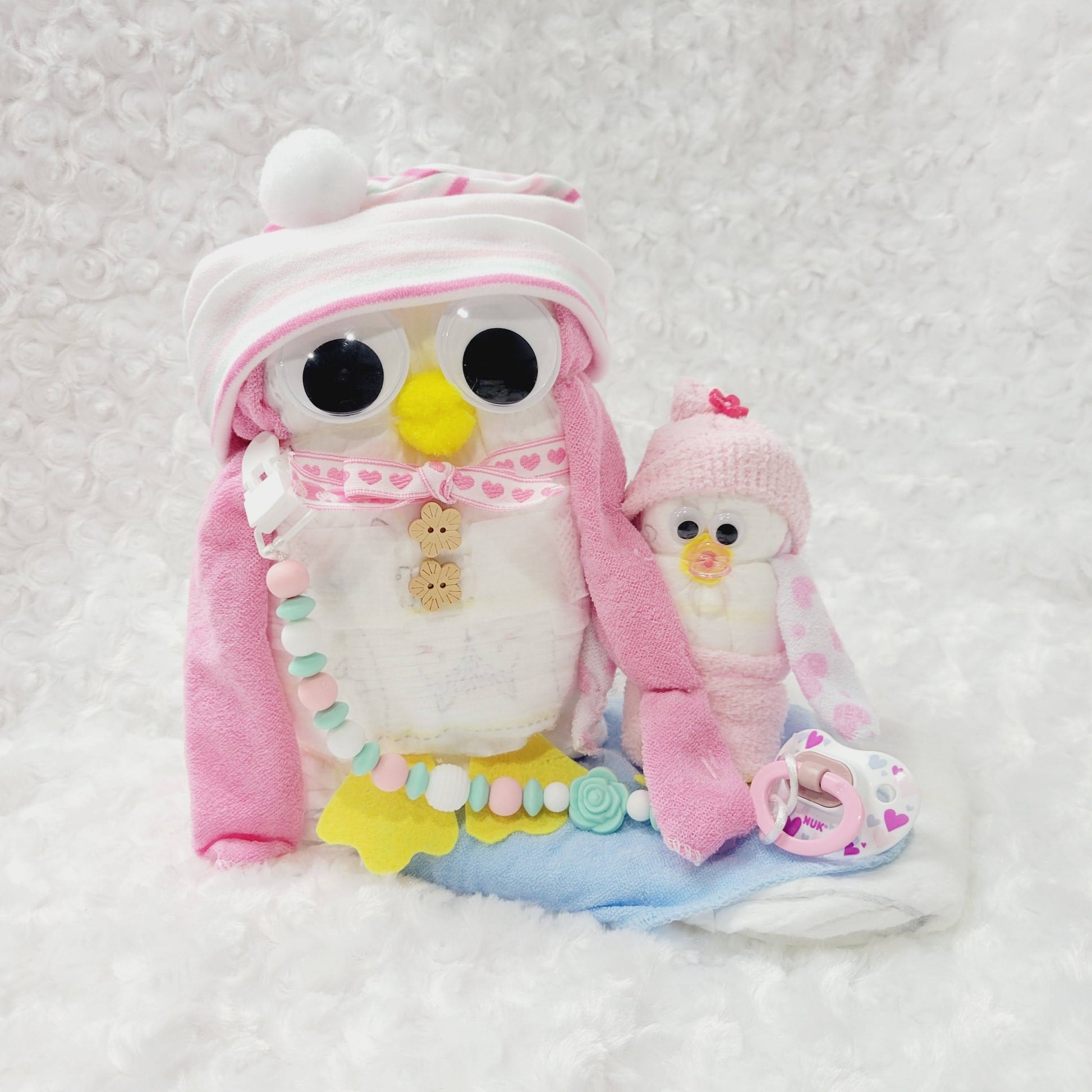 Ensemble cadeau pour babyshower, Couches pour bébé disposé pour former des pingouins rose avec un bonnet en coton, une attache-suce et une suce. 