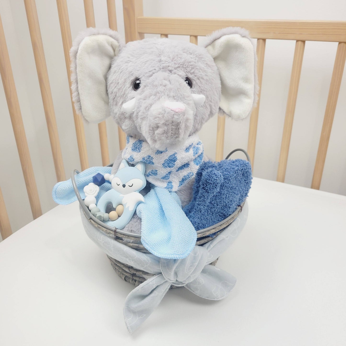Ensemble cadeau our babyshower dans un panier gris contenant un toutou éléphant gris, des débarbouillettes bleu une doudou grise et un jouet de dentition avec un renard bleu.