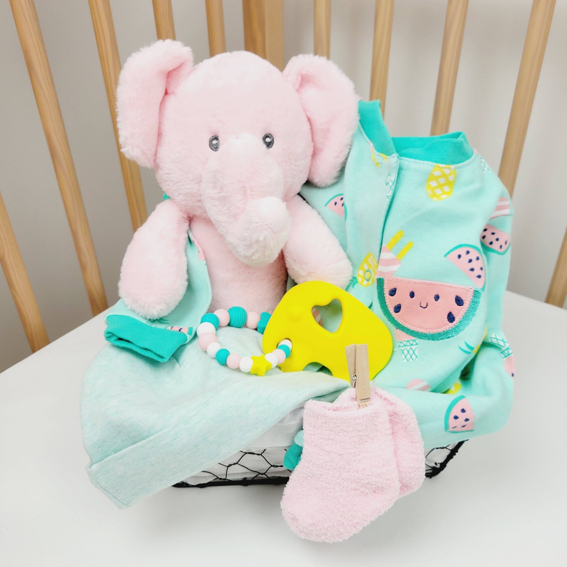 Ensemble cadeau dans un panier pour babyshower. Contient un toutou éléphant rose un pyjamas turquoise avec des melon une paire de bas chaud rose, un bonnet turquoise  et un hochet de dentition en silicone avec un éléphant jaune. 