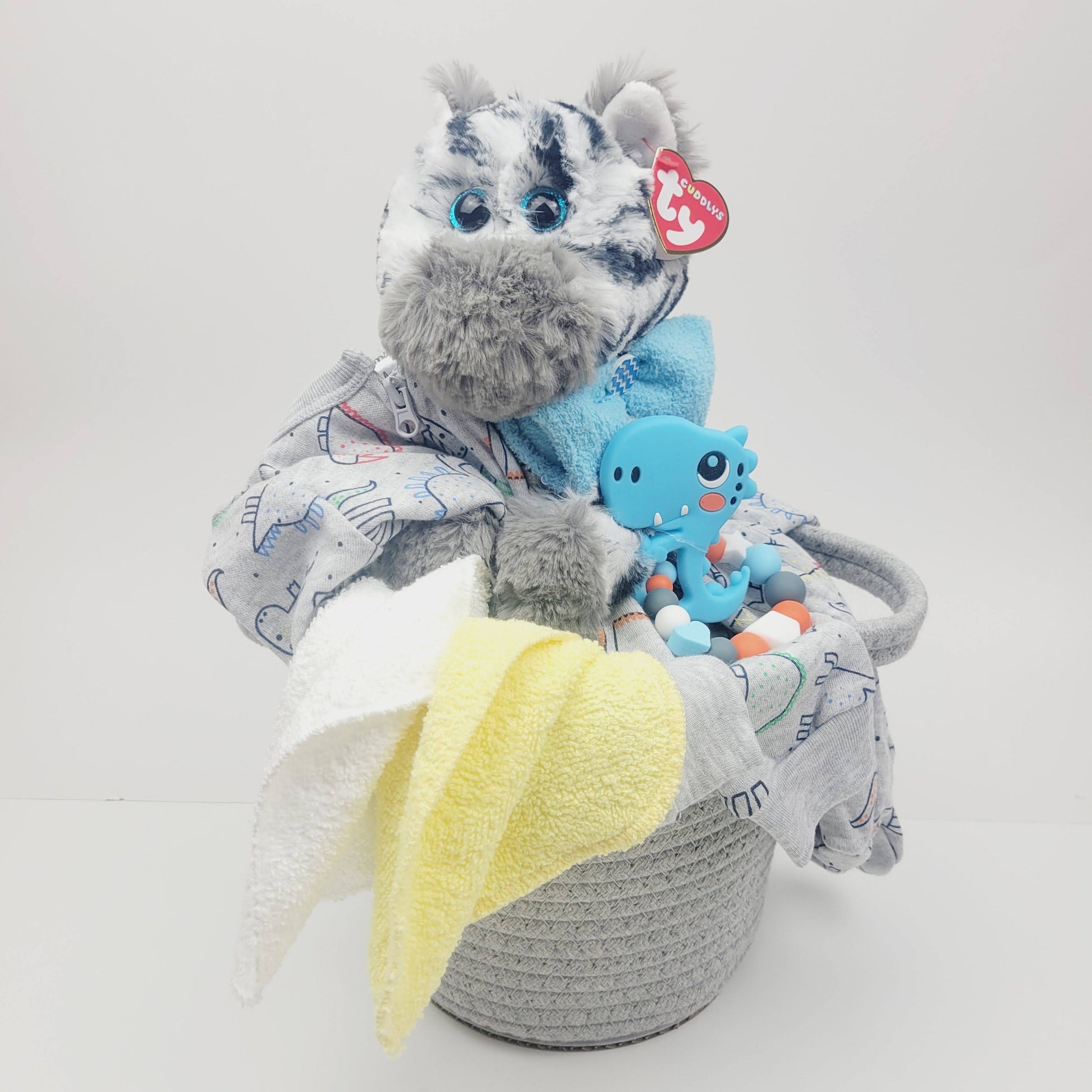 Ensemble cadeau pour babyshower pour bébé. Dans un panier gris on retrouve un toutou zèbre avec un pyjama gris avec des dinosaures, un jouet de dentition avec un dino  bleu. 