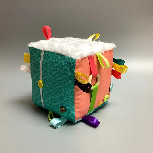 Cube d'éveil pour enfant  Fait en coton avec rubans et petits accessoires de bois. Le cube fait du bruit lorsqu'il est agité.  Ce produit unique est fait au Québec.